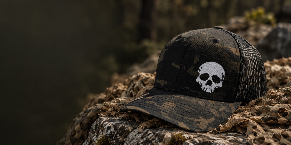 Black Multicam mesh-back cap with a white skull design on coyote netting.